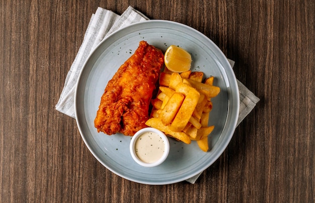 Pesce fritto tradizionale e patatine fritte con salsa tartara in piatto bianco su fondo di legno scuro vista dall'alto