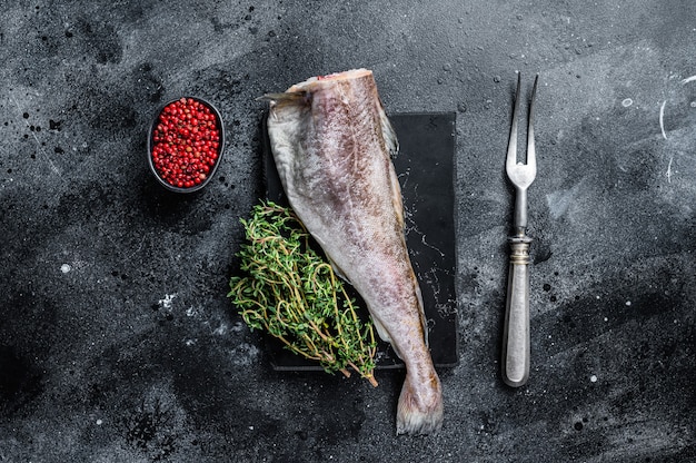 Pesce intero crudo di merluzzo crudo su tavola di marmo con timo. Sfondo nero. Vista dall'alto.