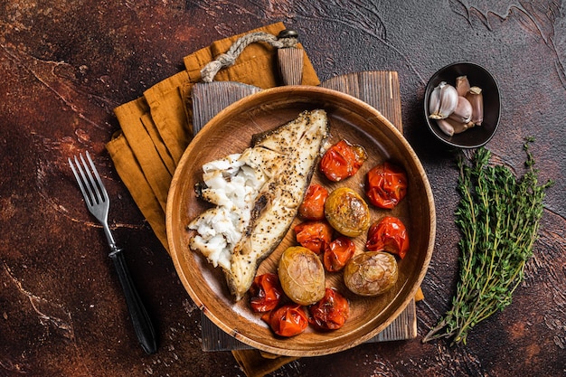 Pesce ippoglosso al forno con pomodoro arrosto e patate nel piatto di legno. Sfondo scuro. Vista dall'alto.