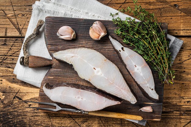 Pesce ippoglosso atlantico, bistecche crude su tavola di legno con erbe aromatiche. Fondo in legno. Vista dall'alto.