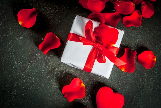 petali di fiori di rosa, confezione regalo incartata bianca con nastro rosso e candela rossa festiva su pietra scura