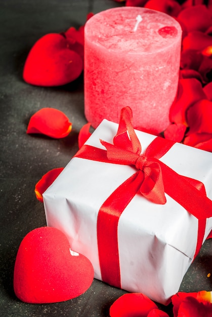 petali di fiori di rosa, confezione regalo incartata bianca con nastro rosso e candela rossa festiva su pietra scura