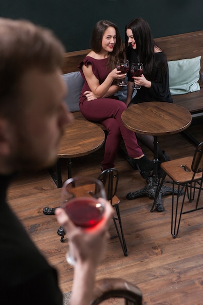 Pettegolezzi sul maschio. Seduzione femminile. Donne civettuole nel bar a fuoco sullo sfondo, uomo irriconoscibile con vino, concetto di flirt