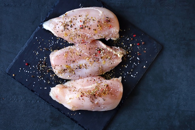 Petti di pollo crudi con spezie e sale marino su un bordo di pietra.