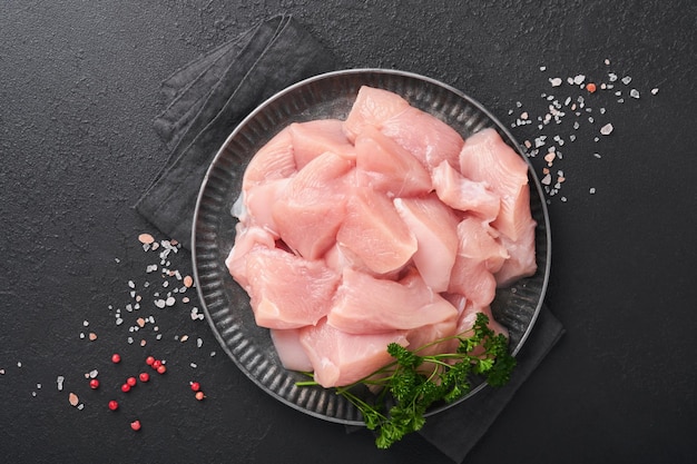 Petto di pollo crudo affettato o tagliato a pezzi su tagliere di legno con erbe e spezie su sfondo di cemento di pietra ardesia scura Carne di pollo cruda Vista dall'alto con spazio per la copia Mock up