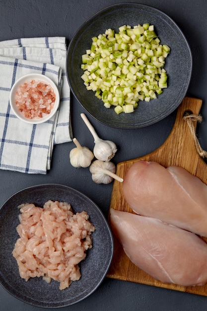 Petto di pollo tritato filetto di pollame su un tagliere e in una ciotola zucchine tritate in una ciotola aglio e sale rosa su un canovaccio su un tavolo di cemento scuro Fare le polpette