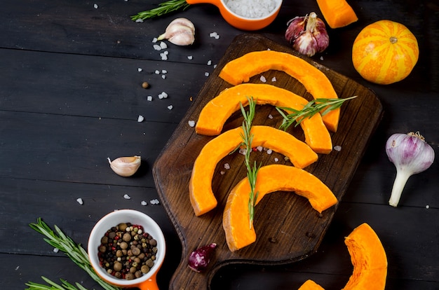 Pezzi crudi di zucca su un tagliere con rosmarino, sale, spezie sul vecchio tavolo scuro. Verdura preparata per la cottura in forno. Cucina casalinga autunnale