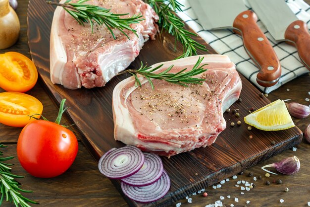 Pezzi di bistecca di maiale cruda con spezie ed erbe aromatiche rosmarino, sale e pepe su fondo di legno in stile rustico.