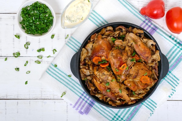 Pezzi di coniglio fritto con funghi di bosco su una padella in ghisa su fondo di legno bianco.