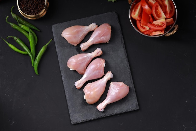 Pezzi di coscia o coscia di pollo crudi disposti su un foglio di grafite con ingredienti da cucina posizionati nelle vicinanze.