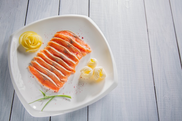 Pezzi di filetto di salmone crudo servito con spezie, limone, burro ed erbe sul piatto bianco e tavolo in legno