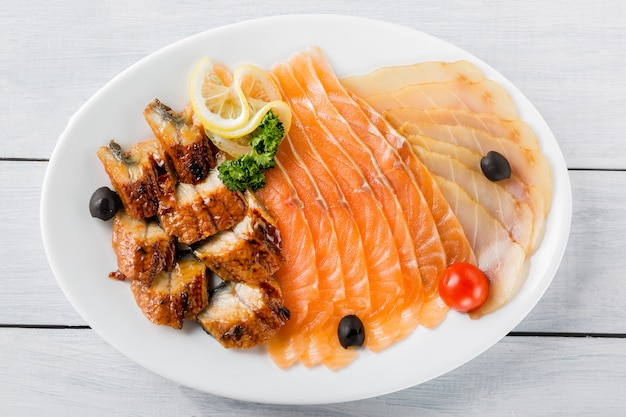 Pezzi di filetto di salmone, grongo, pezzi di storione serviti con limone, olive nere, erbe e pomodoro ciliegia sul piatto bianco e tavolo in legno