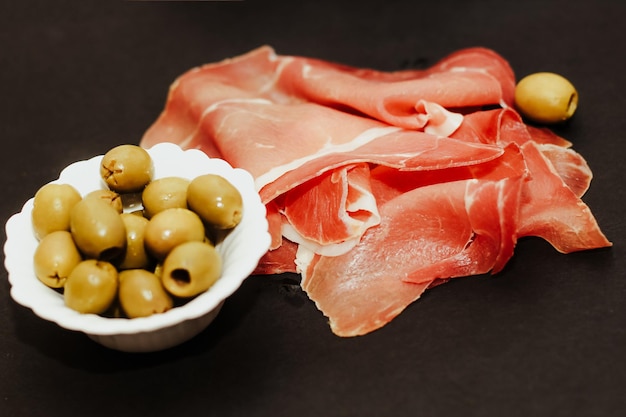 Pezzi di prosciutto di maiale essiccato con olive su una tavola nera