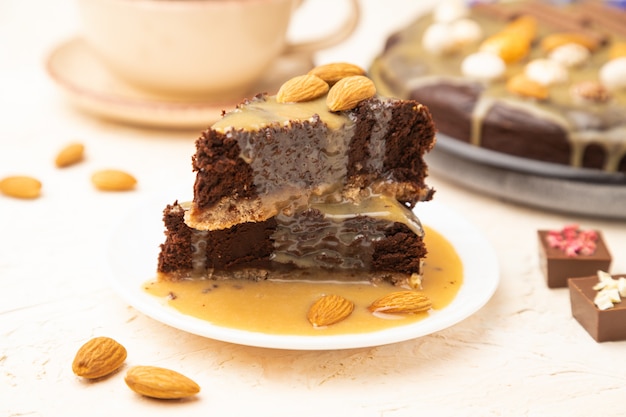 Pezzi di torta brownie al cioccolato con crema al caramello e mandorle su uno sfondo di cemento bianco.