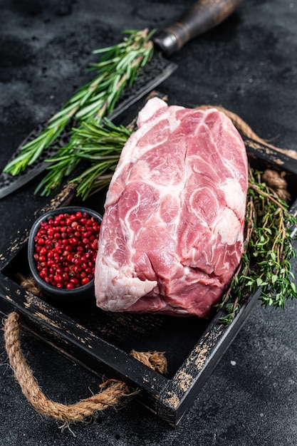 Pezzo di carne di collo di maiale crudo per tagliare la bistecca in vassoio di legno con erbe aromatiche. Sfondo nero. Vista dall'alto.