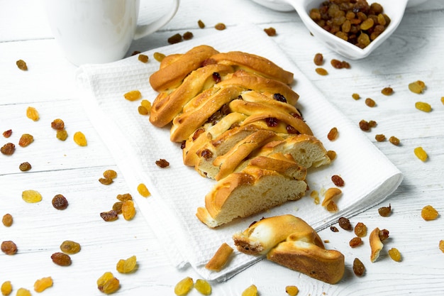 Pezzo di pane intrecciato dolce con l'uva passa sull'asciugamano di cucina su fondo di legno bianco.