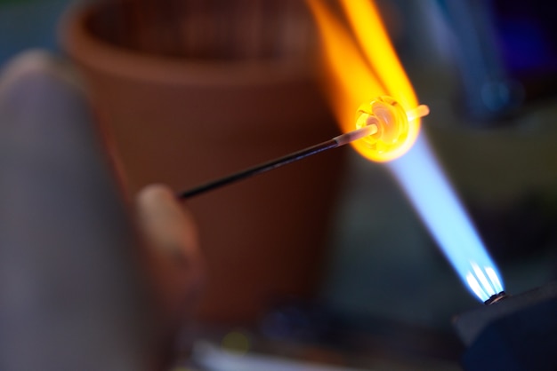 Pezzo di vetro di forma rotonda messo su un bastone ignifugo tenuto da un artigiano sul flusso di fuoco che esce dal bruciatore durante la lavorazione a lume