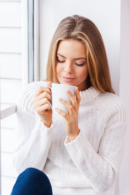 Piacere caldo. Bella giovane donna seduta vicino alla finestra del suo appartamento con in mano una tazza di caffè