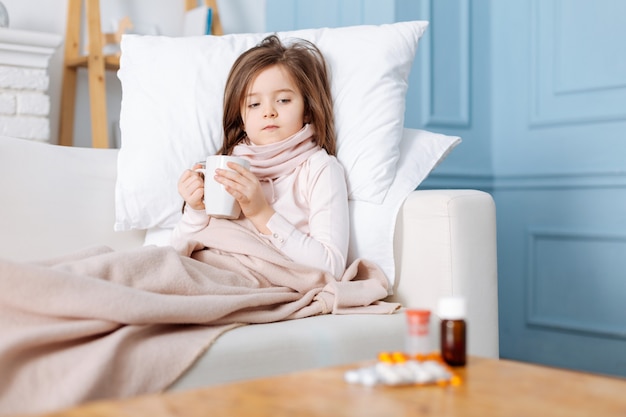 Piacevole bambina malata sdraiata a letto e bere il tè mentre guarda le medicine