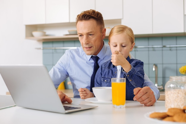 Piacevole bell'uomo bello seduto al tavolo della cucina e utilizzando un laptop mentre ha sua figlia seduta sulle sue ginocchia