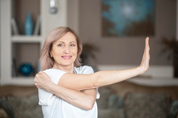 Piacevole donna di mezza età con i capelli biondi che allungano la mano sinistra. Fare yoga a casa