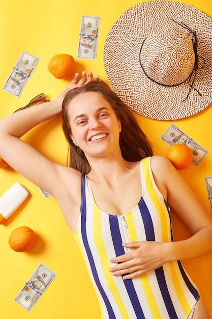 Piacevole donna rilassata sorridente che indossa un costume da bagno a righe sdraiato con crema solare di arance e soldi su sfondo giallo che si gode il suo riposo sembra felice e soddisfatta