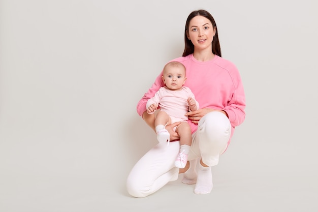 Piacevole giovane donna che indossa abiti casual squat con bambina sulla gamba e guardando direttamente la fotocamera, attraente madre con sua figlia isolata sopra il muro bianco.