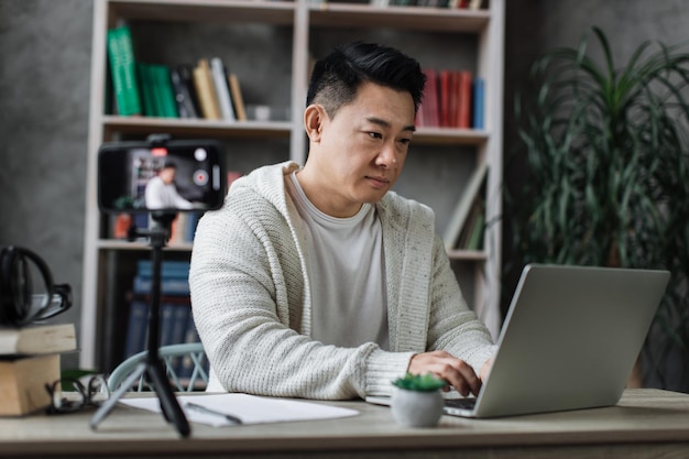Piacevole giovane uomo asiatico che lavora al computer portatile durante la registrazione di video sul suo smartphone sul treppiede