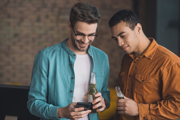 Piacevole uomo felice che tiene il suo smartphone mentre beve birra con il suo amico