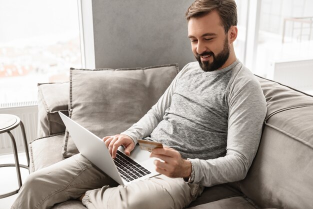 Piacevole uomo moderno anni '30 in abbigliamento casual seduto sul divano nel soggiorno e facendo pagamento con carta di credito e notebook