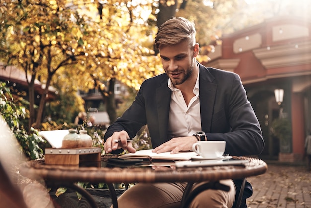 Pianificare la giornata. Bello giovane uomo sorridente in abbigliamento casual intelligente che controlla i suoi appunti nell'agenda personale mentre è seduto al ristorante all'aperto