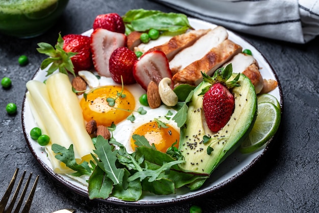 Piastra con un alimento dietetico chetogenico. Uovo fritto, avocado, fragola, filetto di pollo alla griglia, formaggio, noci e rucola. Colazione cheto. Sfondo di ricetta alimentare. Avvicinamento,