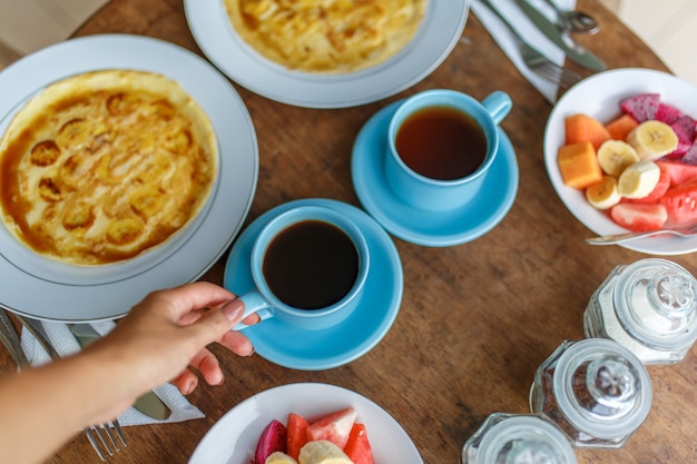 Piatti con pancake alla banana, frutti tropicali e due tazze di caffè sul tavolo di legno.