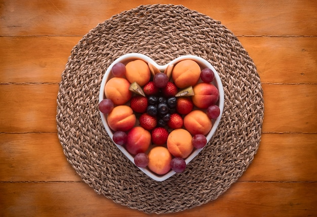 Piatto appetitoso di frutta fresca matura stagionale in un piatto a forma di cuore bianco - mangiare sano con albicocche, uva, fragole, mirtilli e kiwi - Fondo in legno