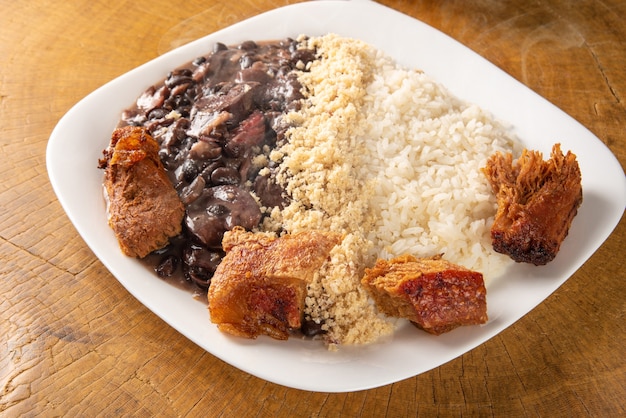 Piatto bianco con una deliziosa feijoada brasiliana, riso, farofa e maiale fritto con due arance su legno rustico