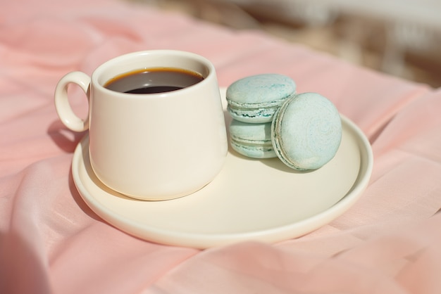 Piatto blu del maccherone francese sul rosa e sulla tazza di caffè che stanno su una tavola di legno con il vaso bianco della tovaglia rosa con le rose e le verdure dei fiori.