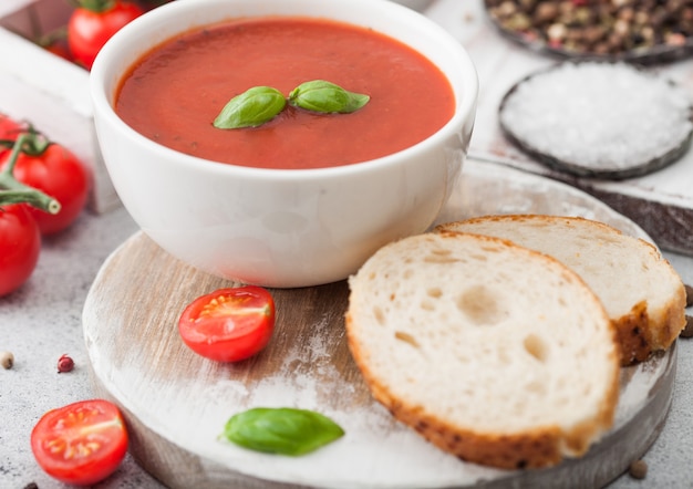 Piatto ciotola bianca di zuppa cremosa di pomodoro con cucchiaio sul tavolo luminoso con scatola di pomodori crudi e pane. Macro