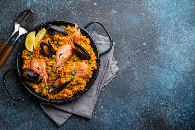 Piatto classico della Spagna, paella di frutti di mare in padella tradizionale su sfondo rustico in cemento blu vista dall'alto. Paella spagnola con gamberi, pinze, cozze, piselli e spicchi di limone fresco dall'alto