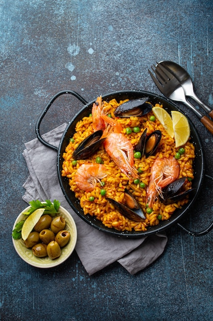 Piatto classico della Spagna, paella di frutti di mare in padella tradizionale su sfondo rustico in cemento blu vista dall'alto. Paella spagnola con gamberi, pinze, cozze, piselli e spicchi di limone fresco dall'alto