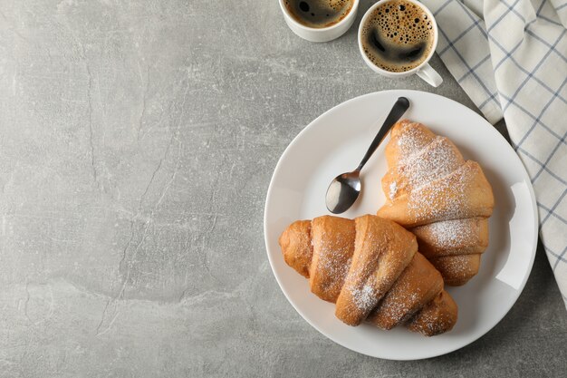 Piatto con i croissant, le tazze di caffè e l'asciugamano su fondo grigio, vista superiore