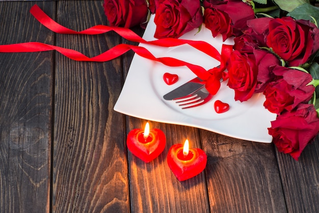 piatto con posate, rose rosse, due candele a forma di cuore e un nastro rosso