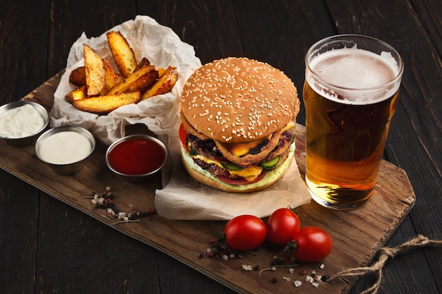 Piatto del ristorante fast food. Hamburger succoso, spicchi di patate, salse e bicchiere di birra fredda su fondo di legno scuro