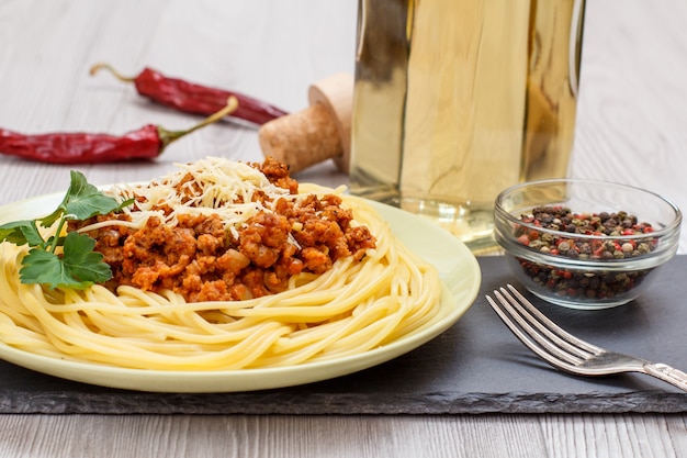 Piatto di porcellana con spaghetti e ragù alla bolognese, ciotola con bacche di pimento, peperoni rossi e bottiglia di vino bianco sullo sfondo.