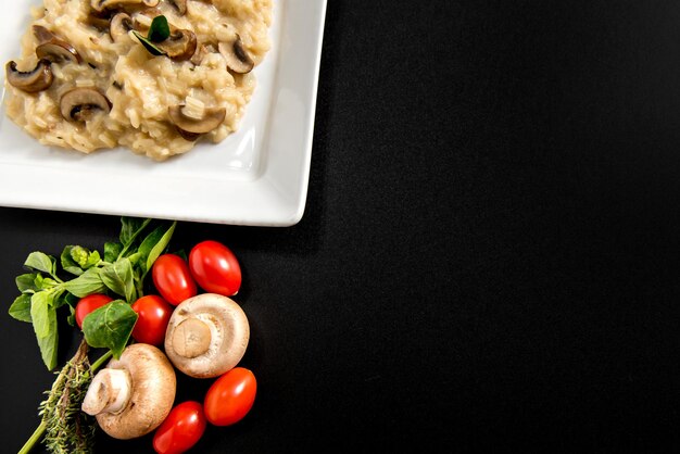 Piatto di risotto ai funghi su sfondo nero pomodorini ed erbe aromatiche su sfondo nero