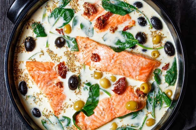 Piatto di salmone toscano di filetto di salmone scottato in padella con salsa di panna, aglio, porro, pomodori secchi, olive e spinaci su una teglia nera su un tavolo di legno scuro, vista dall'alto, primo piano