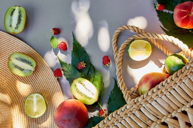 Piatto disteso di cappello di paglia e borsa con frutta lampone, pesca, kiwi, lime su superficie grigia alla luce del sole, ora legale. Concetto di drogheria, copia spazio, vista dall'alto.