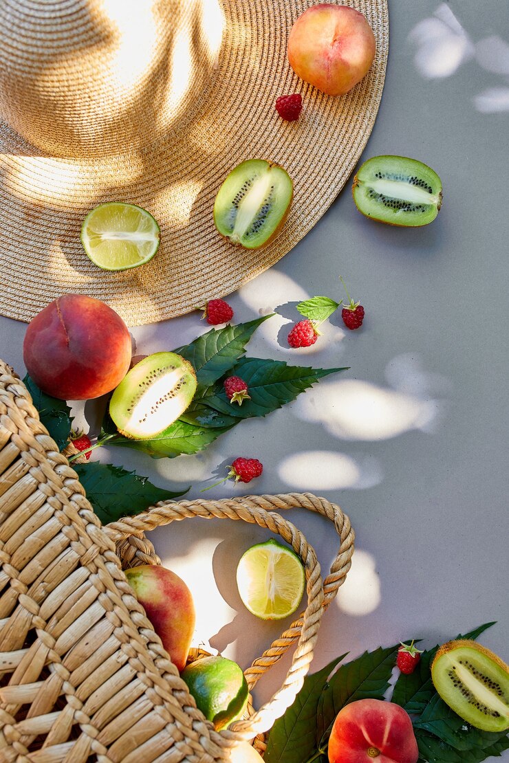 Piatto disteso di cappello di paglia e borsa con frutta lampone, pesca, kiwi, lime su superficie grigia alla luce del sole, ora legale. Concetto di drogheria, copia spazio, vista dall'alto.