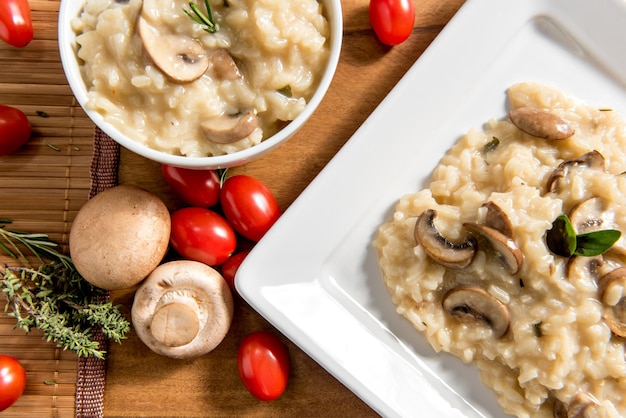 Piatto e ciotola con delizioso risotto ai funghi su tavola di legno pomodoro ciliegino erbe aromatiche e funghi