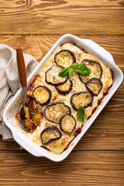 Piatto mediterraneo greco Moussaka con melanzane al forno, carne macinata in casseruola di ceramica bianca con tovagliolo su fondo rustico in legno dall'alto, piatto tradizionale della Grecia