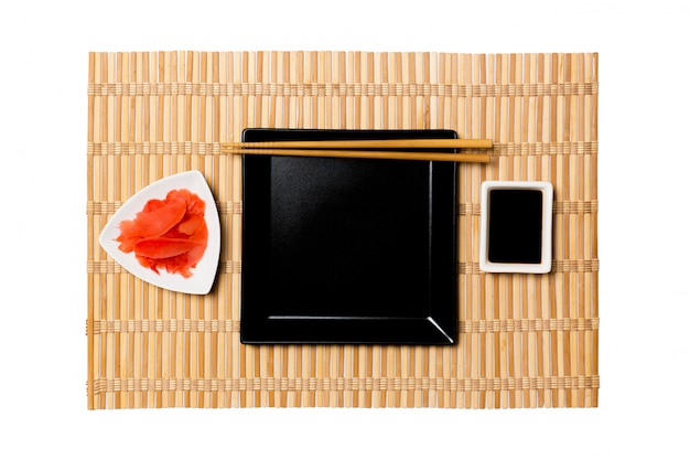 Piatto quadrato nero vuoto con le bacchette per salsa di sushi, zenzero e soia sulla stuoia di bambù giallo